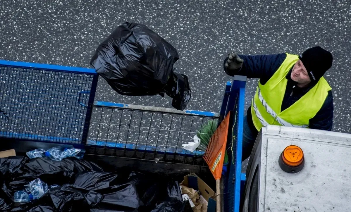 Worker in a reflective vest tossing a black trash bag into a sanitation truck
